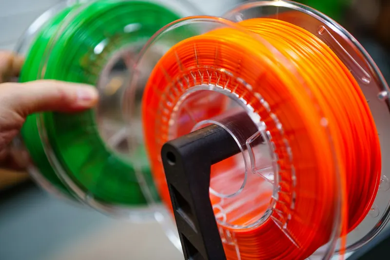 A hand putting a spool of green filament next to a spool of orange filament