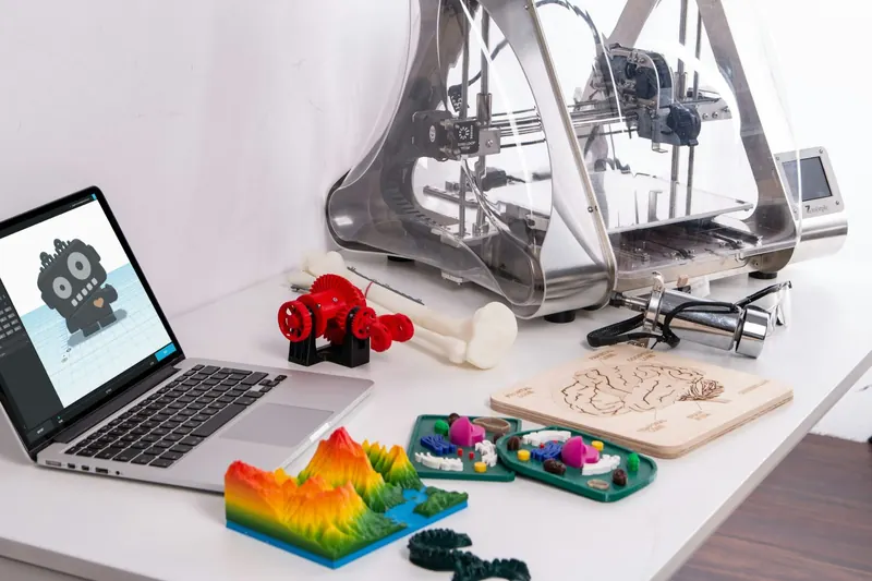 A laptop showing a 3D model, with a 3D printer and colourful prints on a white desk